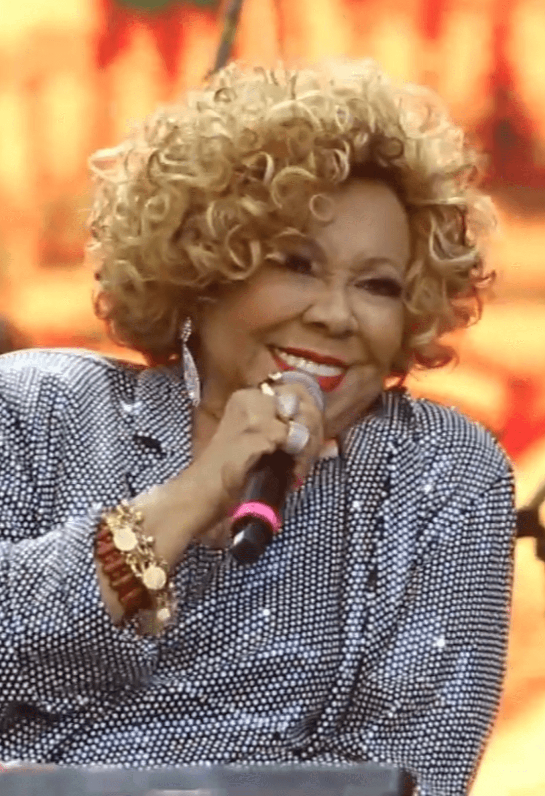 Alcione, de cabelo loiro cacheado e roupa prateada e brilhante, sorri ao microfone