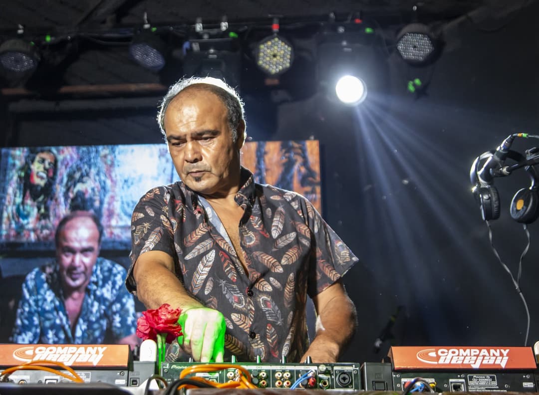 DJ de camisa estampada mexe na mesa de som sob luzes de palco, com um telão ao fundo