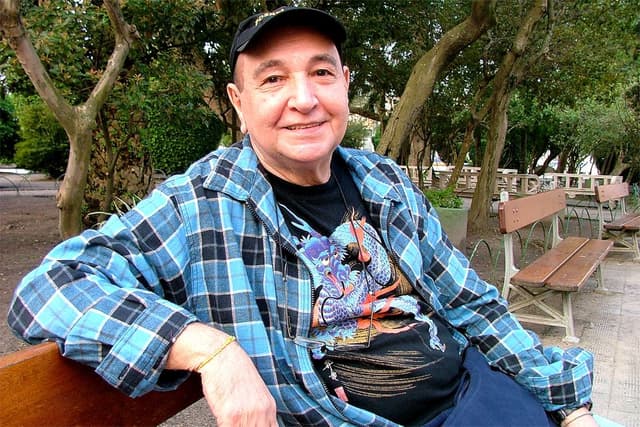 João Donato sorri sentado num banco de praça, de boné e camisa xadrez azul