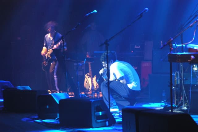 O Mombojó num palco iluminado de azul, com um guitarrista de pé e outro músico agachado junto ao microfone