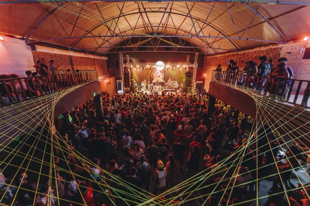 Plateia lotada diante do palco, num galpão de teto em arco cortado por fios de luz verde, no Festival DoSol 2025