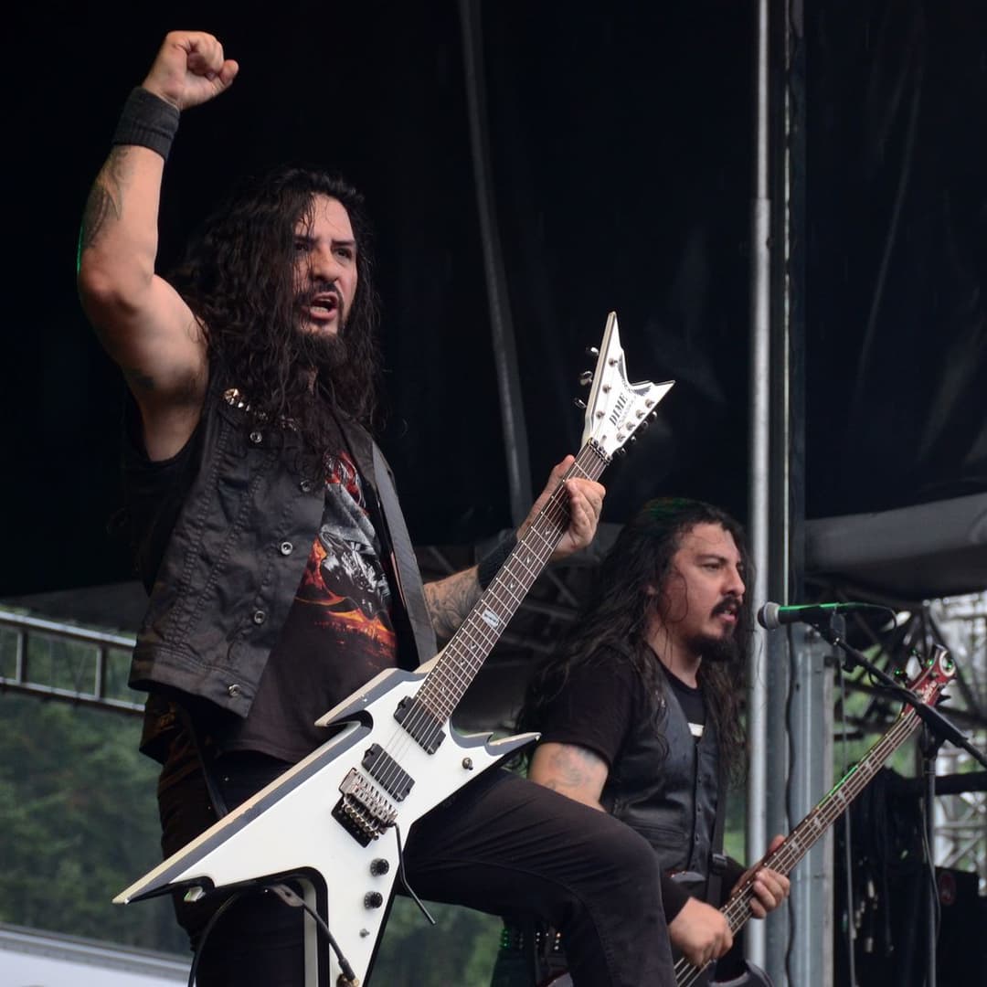 O guitarrista do Krisiun ergue o punho com a guitarra branca ao lado do baixista, que canta ao microfone