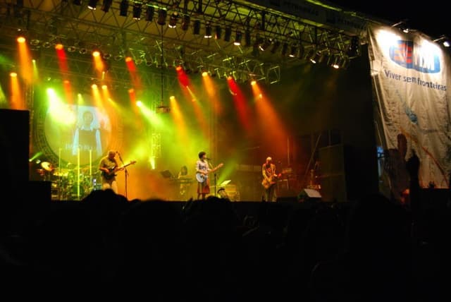 O Pato Fu num palco grande sob luzes coloridas, com o nome da banda no telão ao fundo