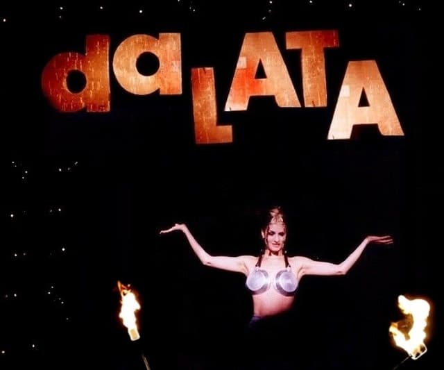 Cantora de braços abertos no palco, sob o letreiro “da lata” e chamas nas laterais