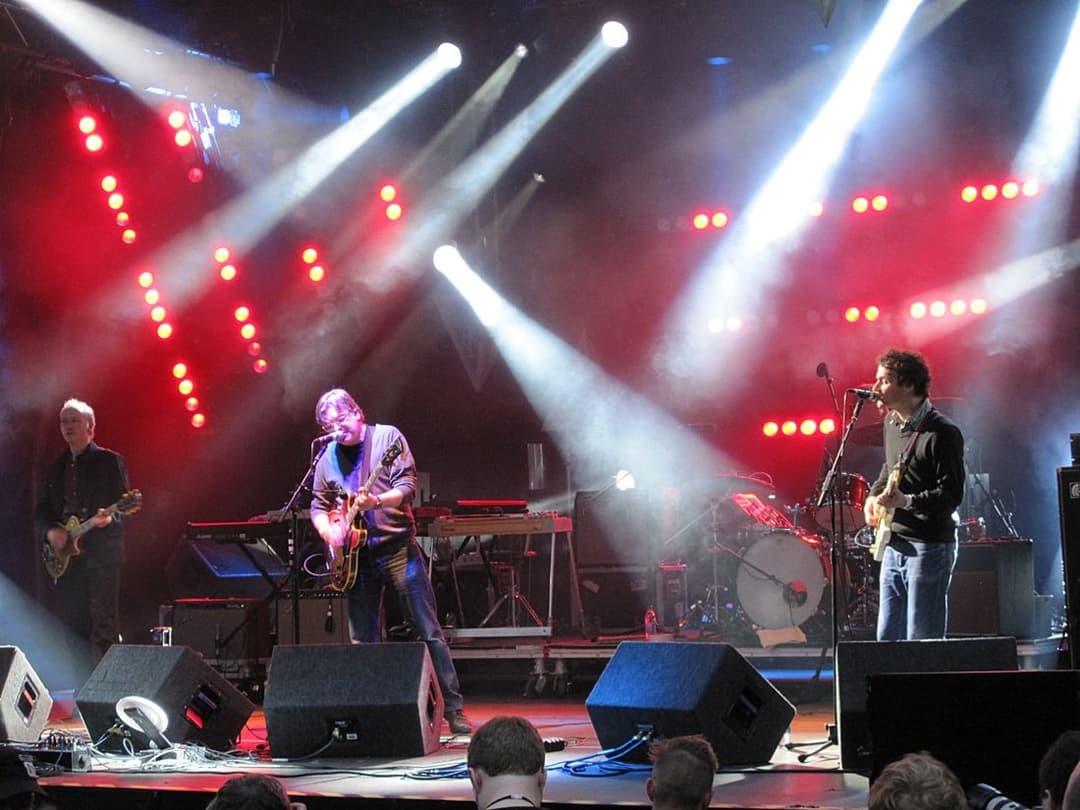 Quatro músicos tocam guitarra e bateria num palco com luzes vermelhas e brancas