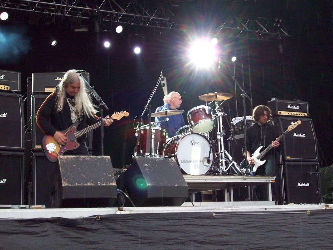 Banda de três integrantes no palco, com o guitarrista de cabelos longos e brancos à esquerda
