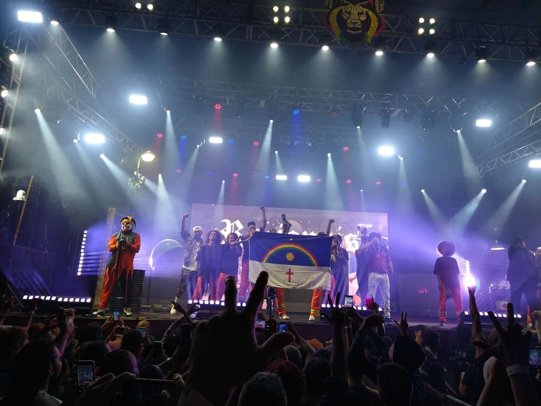 Grupo de rap no palco segura a bandeira de Pernambuco diante da plateia de mãos erguidas