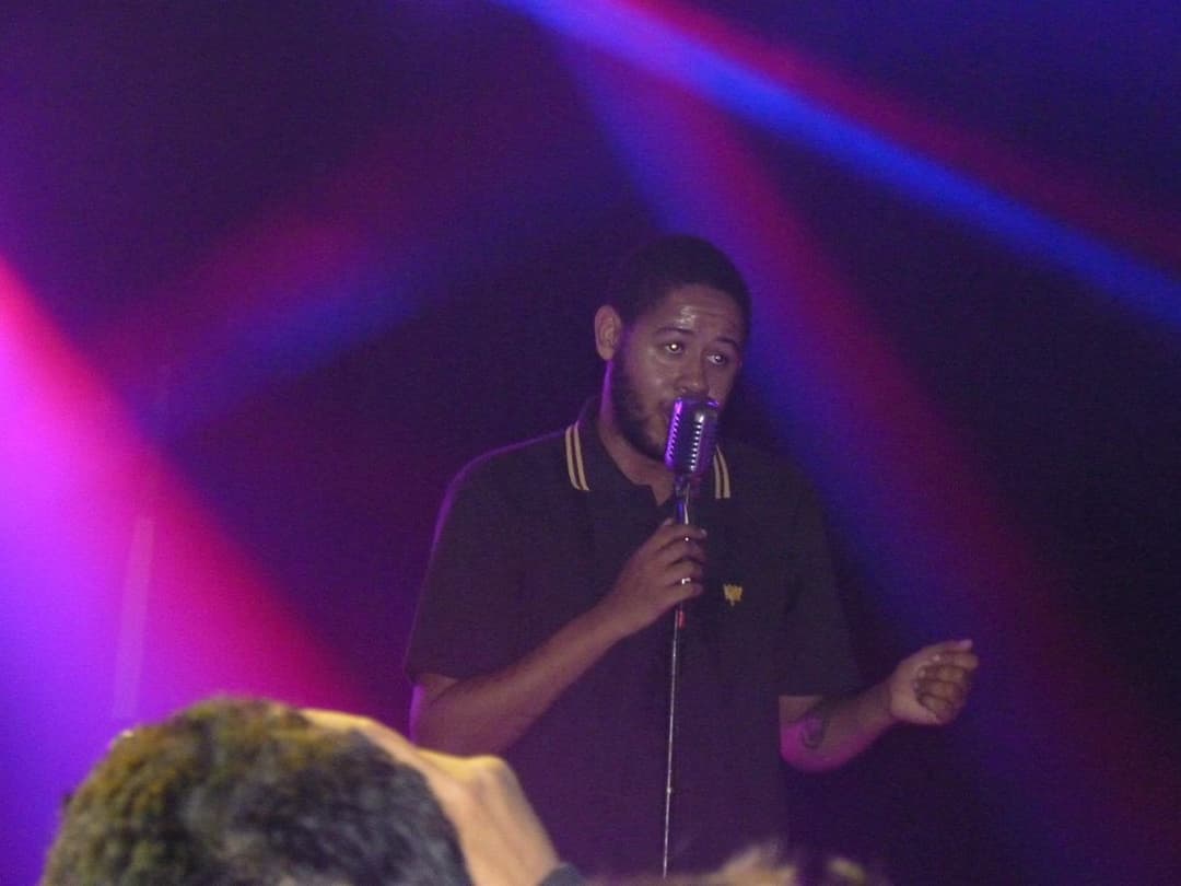 Rapper de camisa polo escura canta ao microfone sob luzes azuis e vermelhas