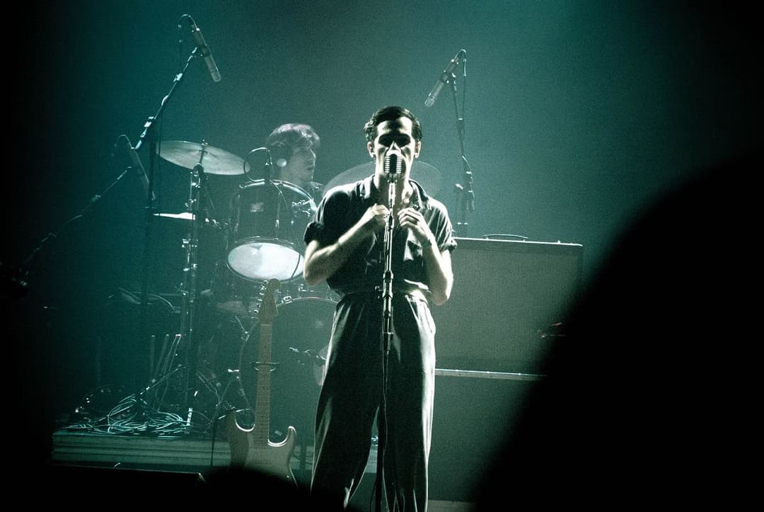 Cantor de camisa escura canta ao microfone num palco iluminado de verde, com o baterista ao fundo