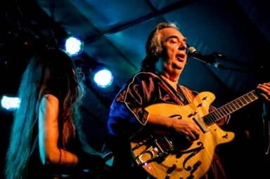 Sérgio Dias canta e toca guitarra no palco, ao lado de uma guitarrista de cabelo comprido