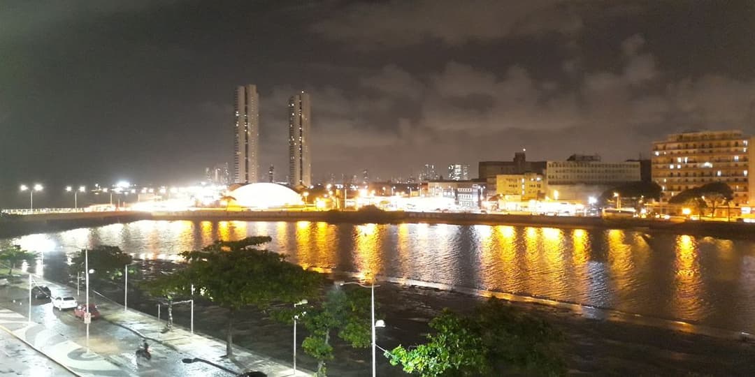 O Rio Capibaribe à noite, com as luzes da outra margem refletidas na água e o cais arborizado em primeiro plano