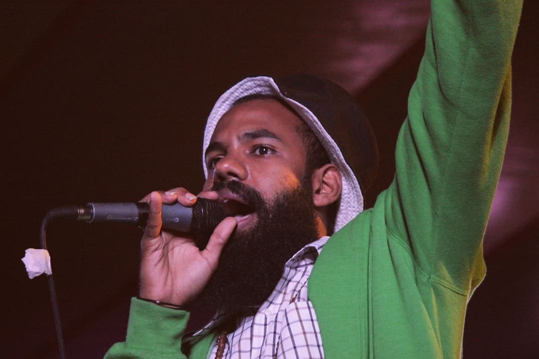 Cantor de barba e chapéu canta ao microfone com o braço erguido