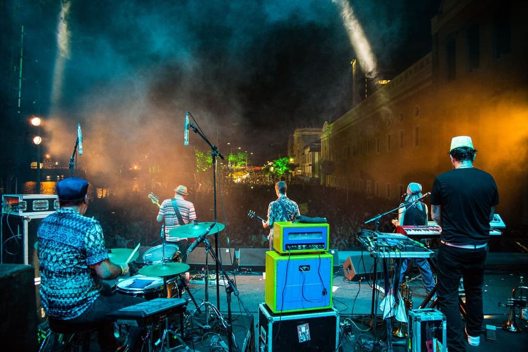 Banda vista de trás do palco, tocando para a multidão, com um casarão iluminado ao fundo
