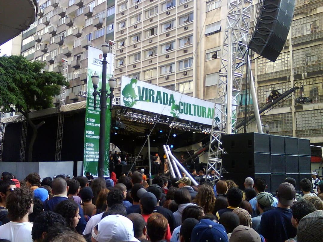 Multidão diante de um palco montado entre prédios, sob a faixa da Virada Cultural