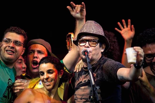 Vocalista de chapéu e óculos canta ao microfone, cercado de gente no palco