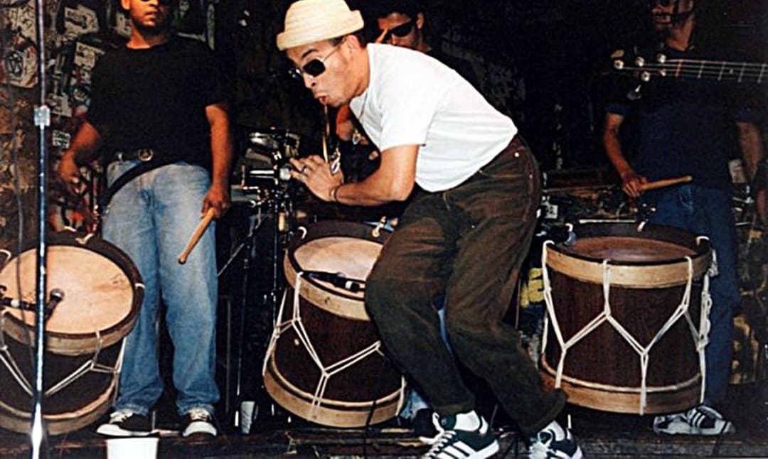 Cantor de boné e óculos escuros se inclina entre tambores de maracatu, no palco