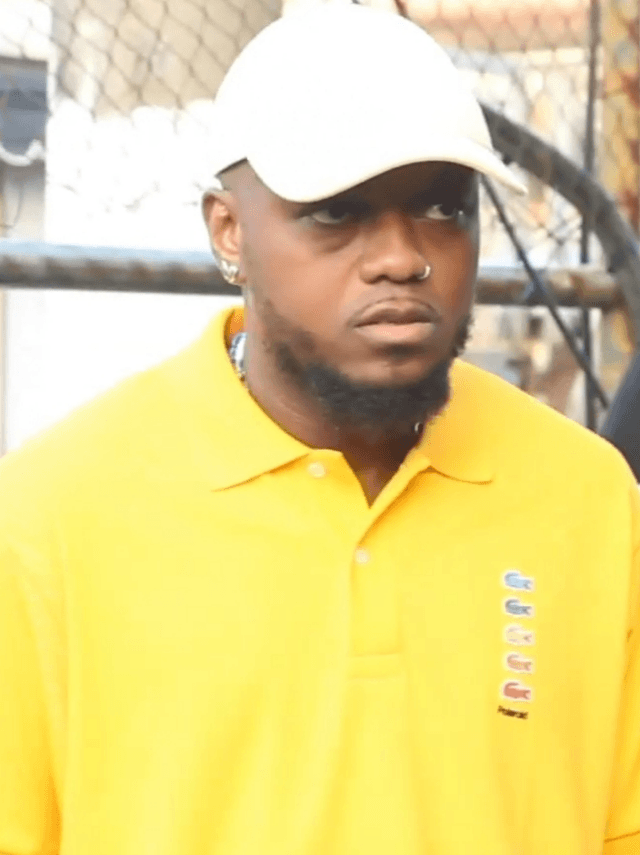 Rapper de boné branco e camisa polo amarela olha para a câmera