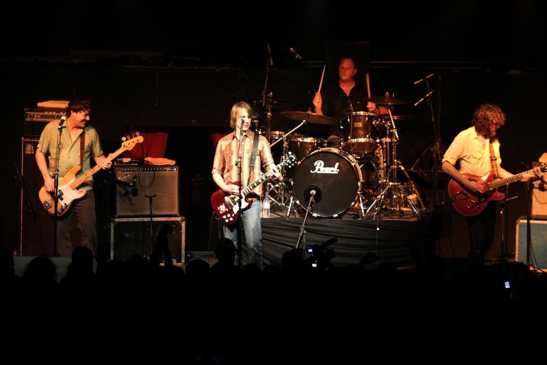 Quatro músicos no palco, com baixo, duas guitarras e bateria, diante das silhuetas da plateia