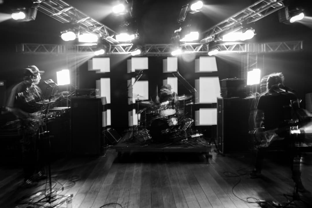 Foto em preto e branco de uma banda tocando num palco com treliças de luz e piso de madeira