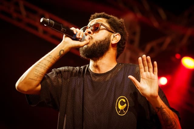 Emicida, de óculos escuros e camiseta preta, canta ao microfone com a mão erguida