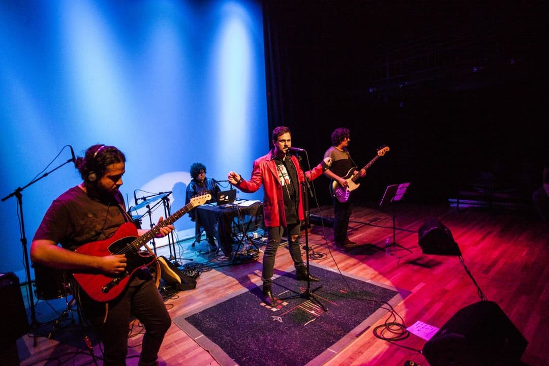Banda toca num palco de teatro com luz azul e vermelha; o vocalista, de jaqueta vermelha, canta ao centro