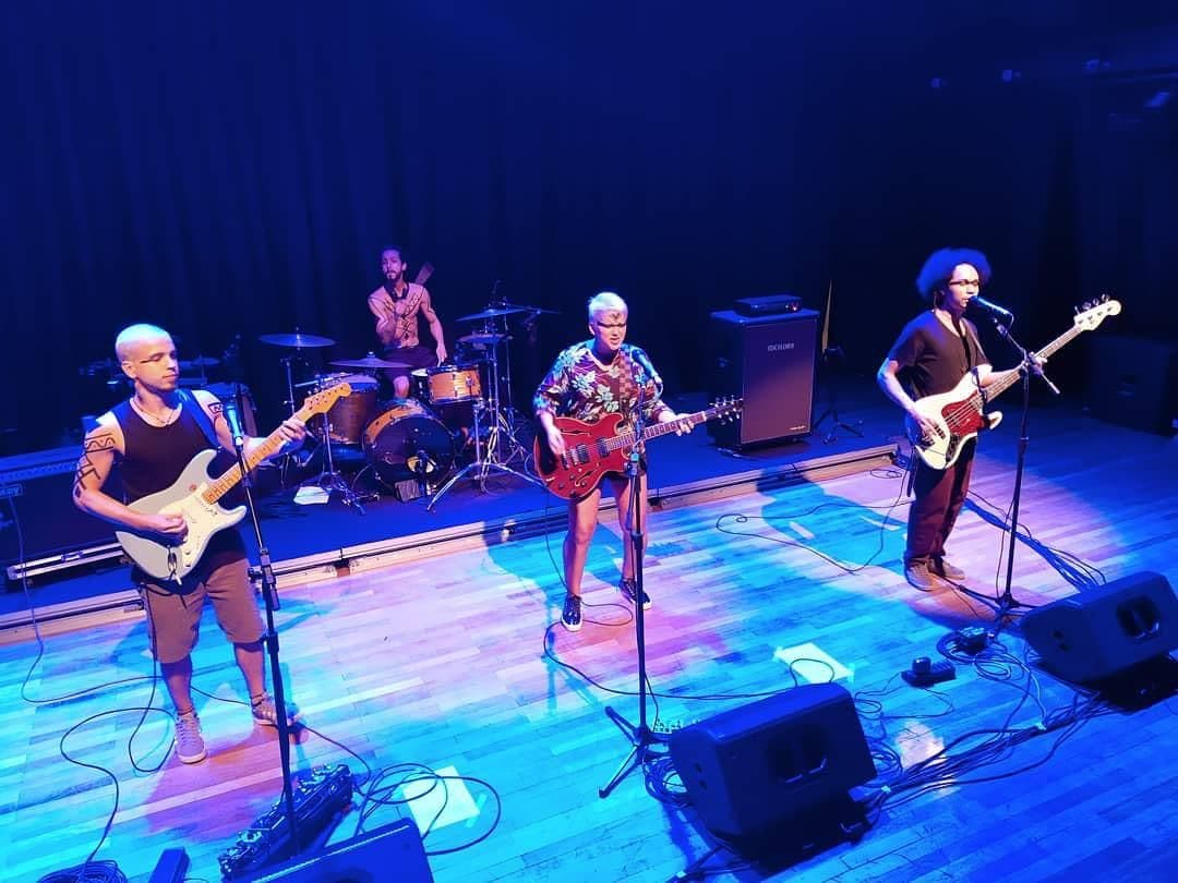 Quatro músicos tocam num palco de teatro com piso de madeira e luz azul