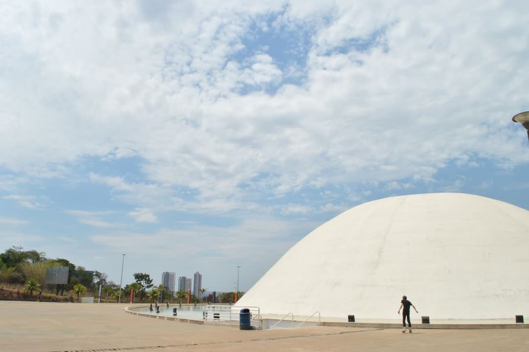 Cúpula branca de concreto sobre uma esplanada, com prédios de Goiânia ao fundo