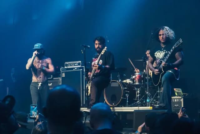 Banda de rock no palco sob luz azul: vocalista sem camisa e de boné, baixista, baterista e guitarrista de cabelo cacheado claro