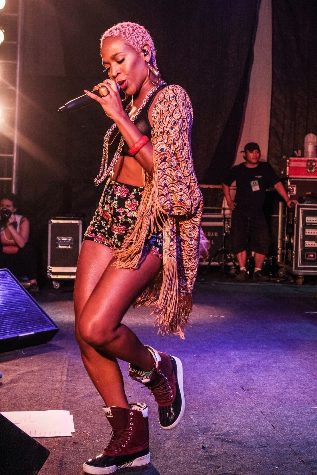 Karol Conká canta num palco de luz lilás, de cabelo trançado claro, kimono de franjas e short estampado