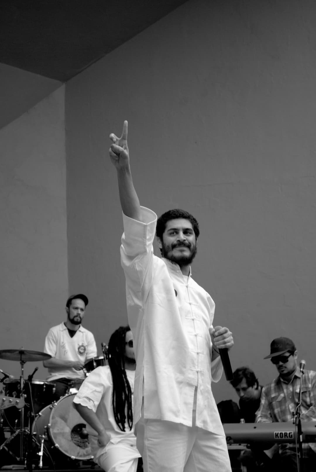 Foto em preto e branco de Criolo, de roupa clara, apontando para o alto com o microfone na mão, à frente da banda