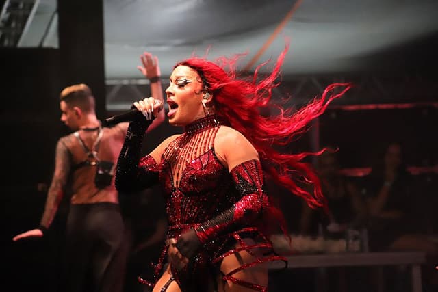Gloria Groove, de cabelo vermelho esvoaçante e roupa de pedrarias, canta ao microfone com um bailarino atrás