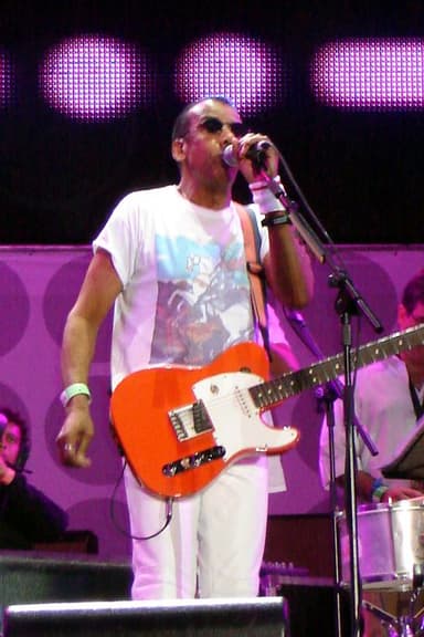 Jorge Ben Jor de camiseta branca canta ao microfone com uma guitarra laranja, diante de um painel roxo