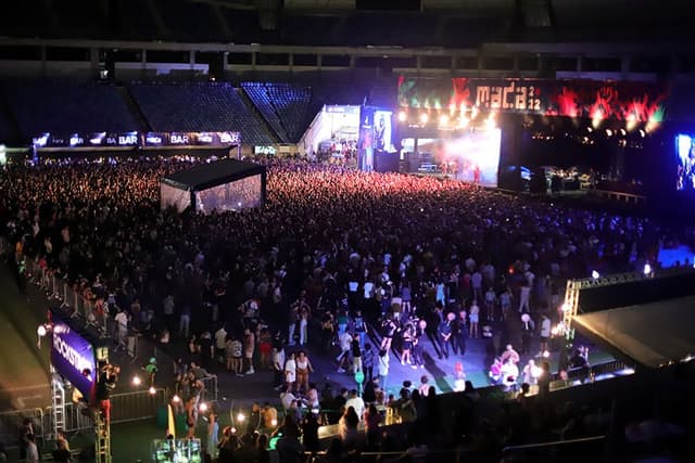 O gramado da Arena das Dunas tomado pelo público à noite, visto do alto, com o palco iluminado ao fundo