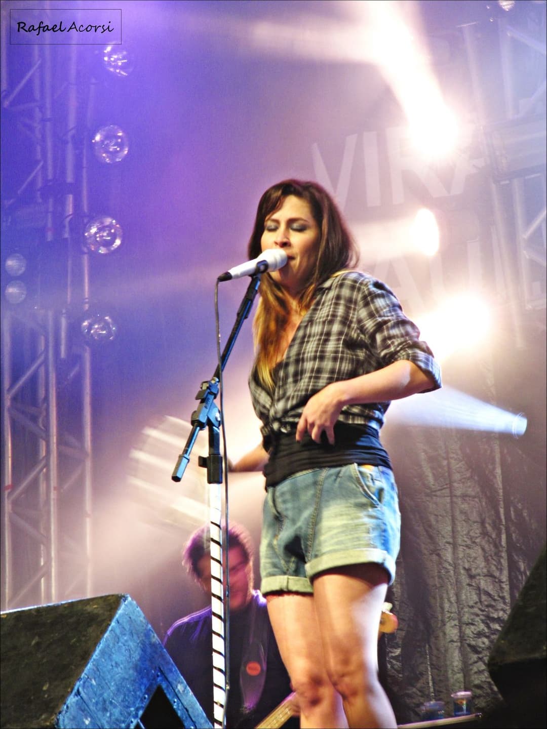 Pitty, de camisa xadrez e short jeans, canta ao microfone num palco iluminado de lilás