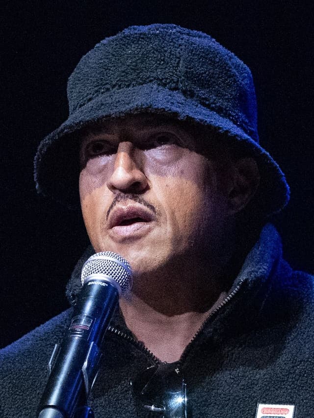 Mano Brown, de chapéu preto felpudo, fala ao microfone diante de um fundo escuro