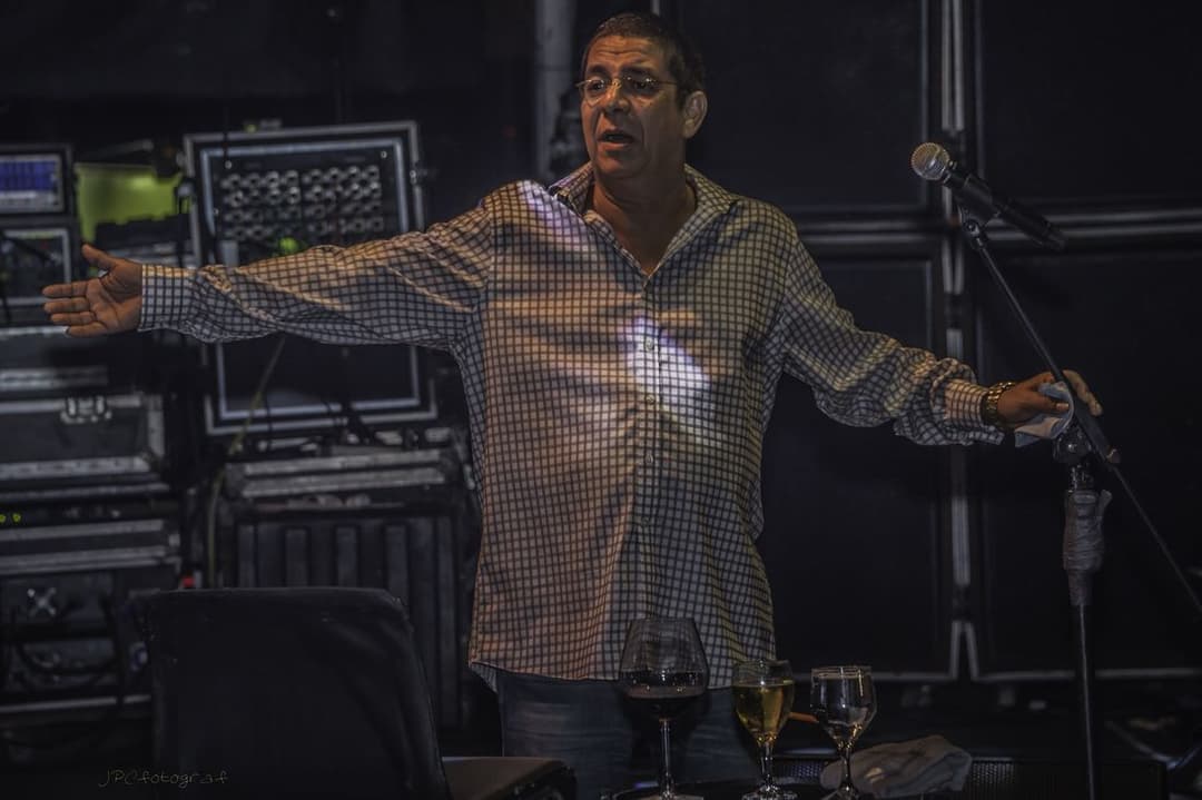 Zeca Pagodinho, de camisa xadrez, abre os braços num palco escuro, entre microfones e amplificadores