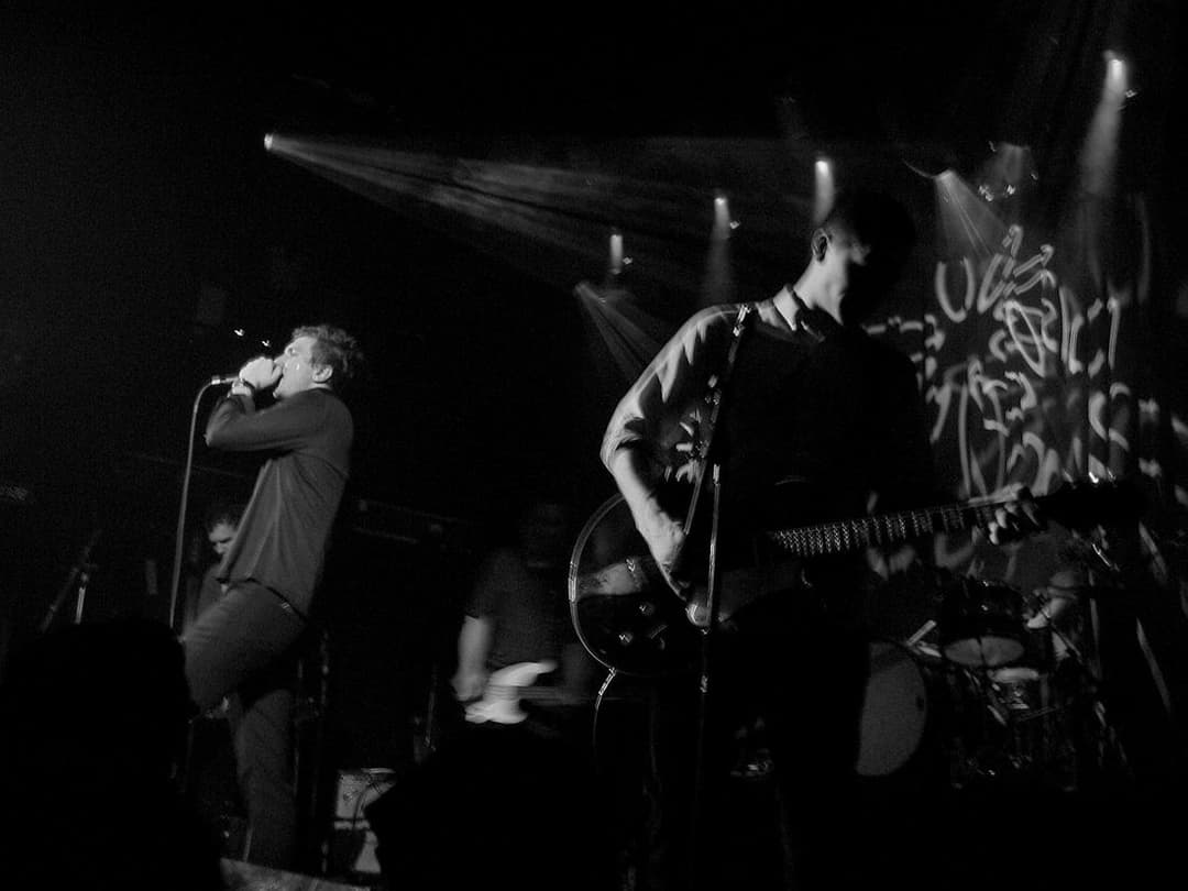 Foto em preto e branco de uma banda num palco escuro: o vocalista canta à esquerda e o guitarrista toca sob um facho de luz