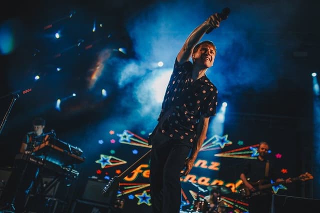 O vocalista do Franz Ferdinand, de camisa escura estampada, estende o braço para a plateia diante de um letreiro luminoso com o nome da banda