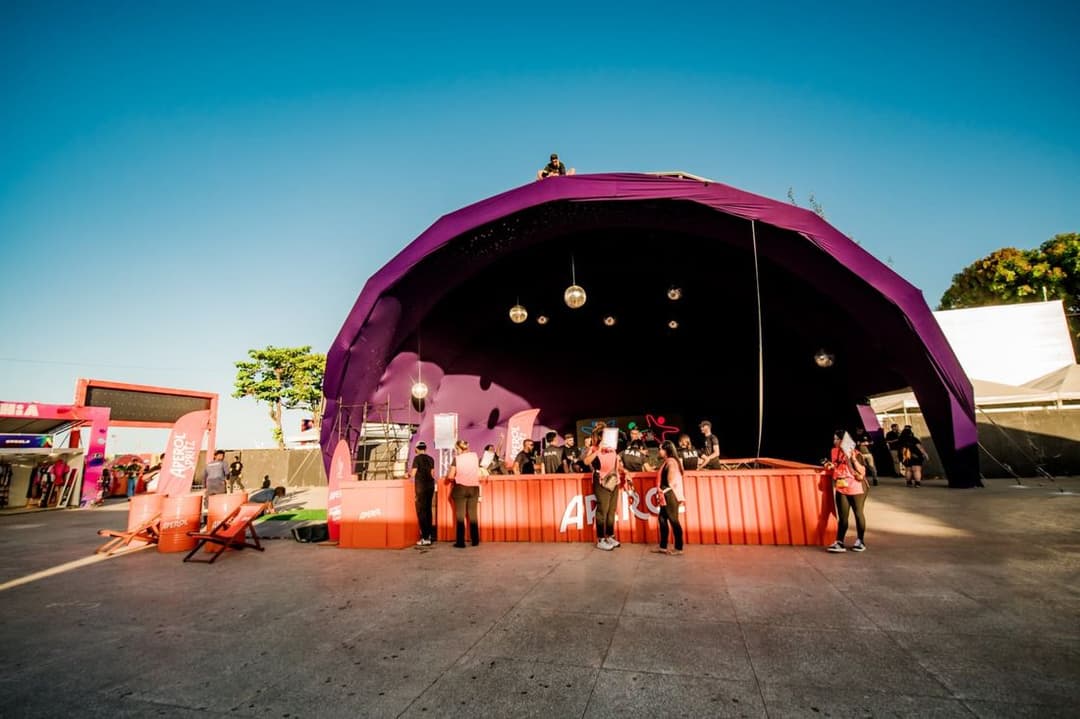 Palco coberto por uma cúpula roxa, com pessoas e barracas em volta, sob céu azul de fim de tarde