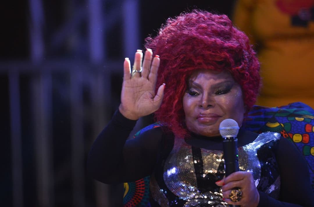 Elza Soares, de cabelo vermelho e roupa colorida e brilhante, acena com a mão erguida enquanto segura o microfone