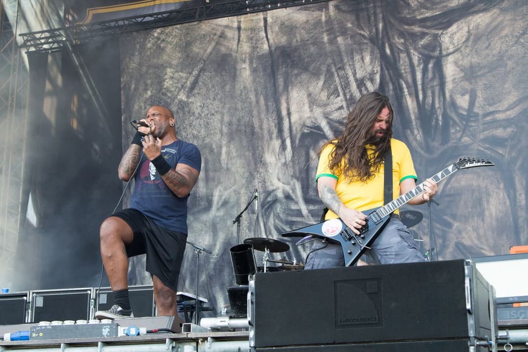 Vocalista careca de regata canta e guitarrista de cabelo comprido e camiseta amarela toca num palco grande ao ar livre