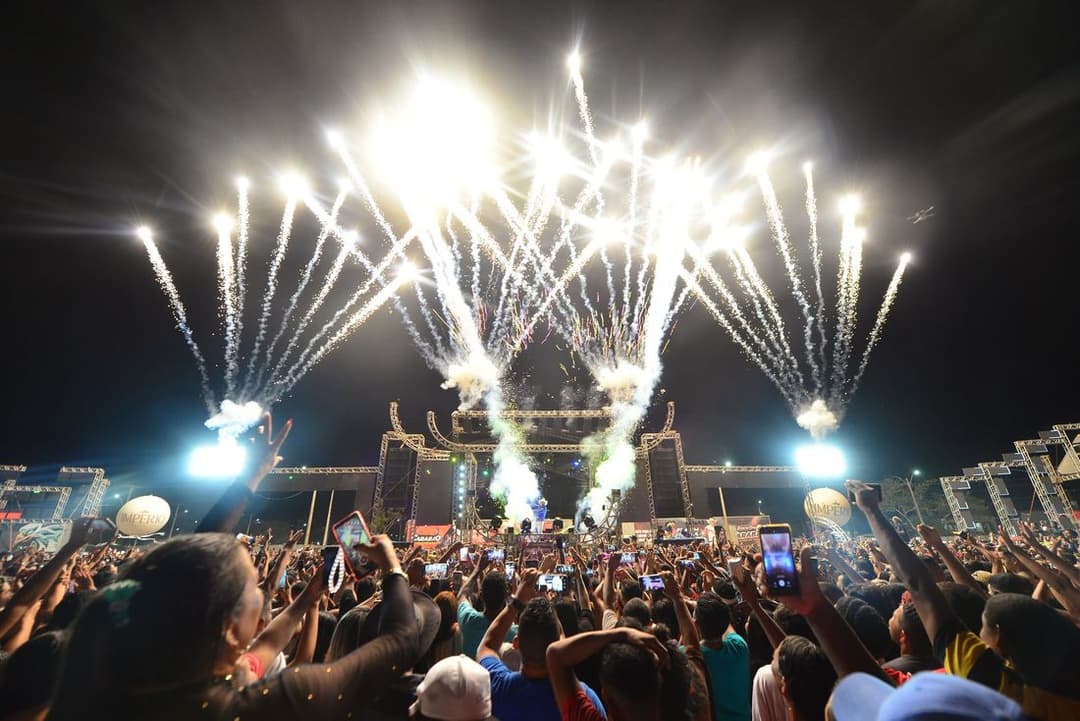 Fogos de artifício explodem no céu noturno sobre um palco iluminado, diante de uma multidão