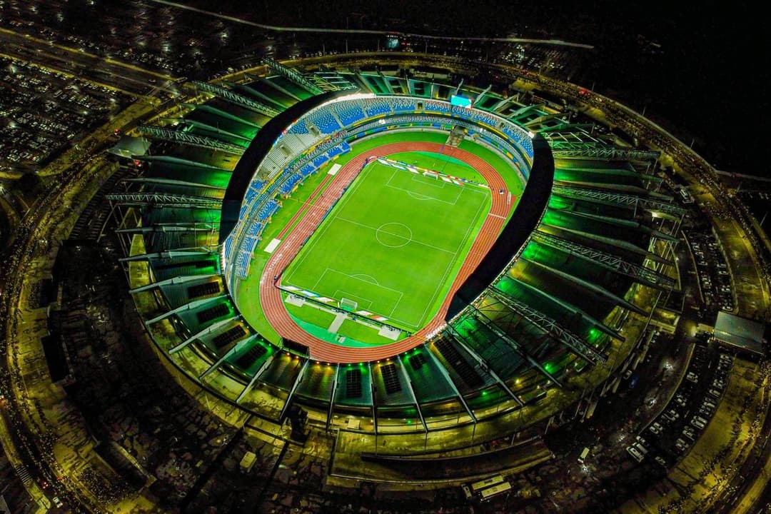 Vista aérea noturna de um estádio oval iluminado, com gramado verde e arquibancadas cercadas de árvores
