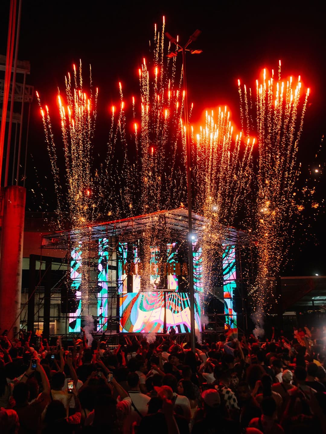 Fogos de artifício sobem atrás de um palco com telão colorido, diante de uma multidão com celulares erguidos à noite