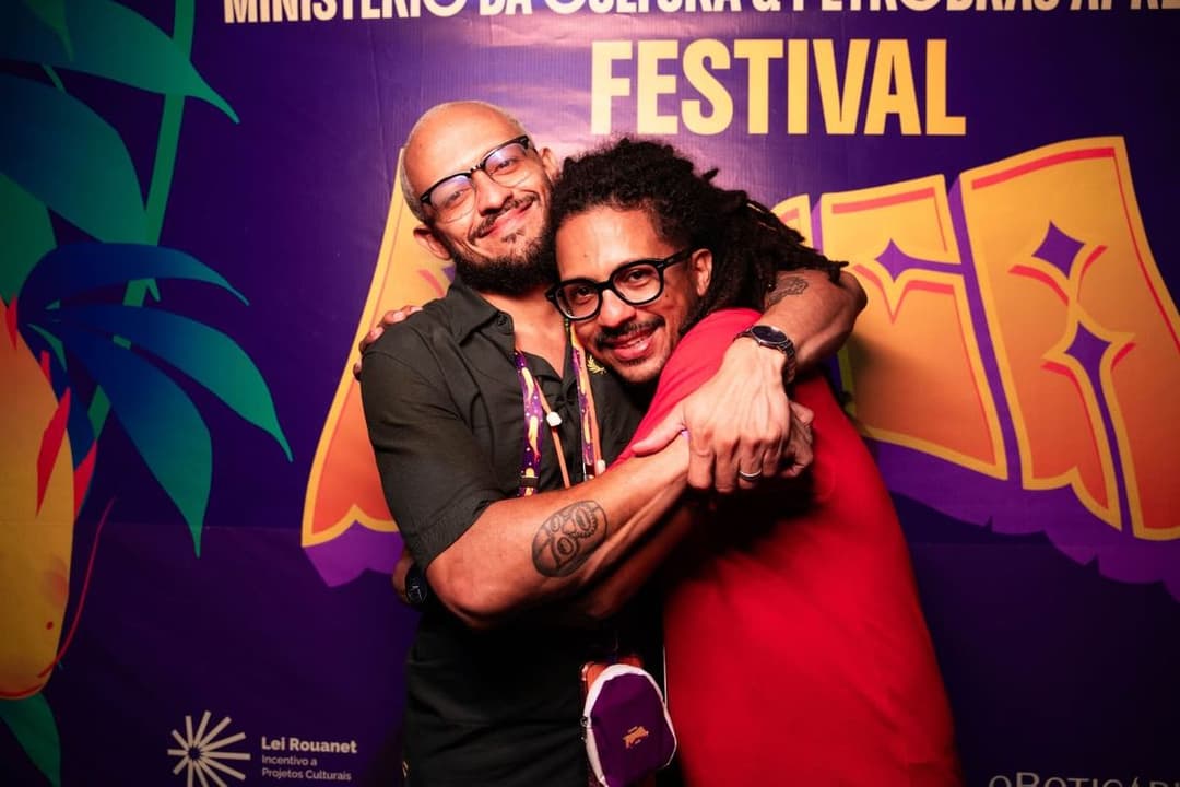 Dois homens de óculos se abraçam sorrindo diante de um painel roxo com folhagens e a palavra Festival