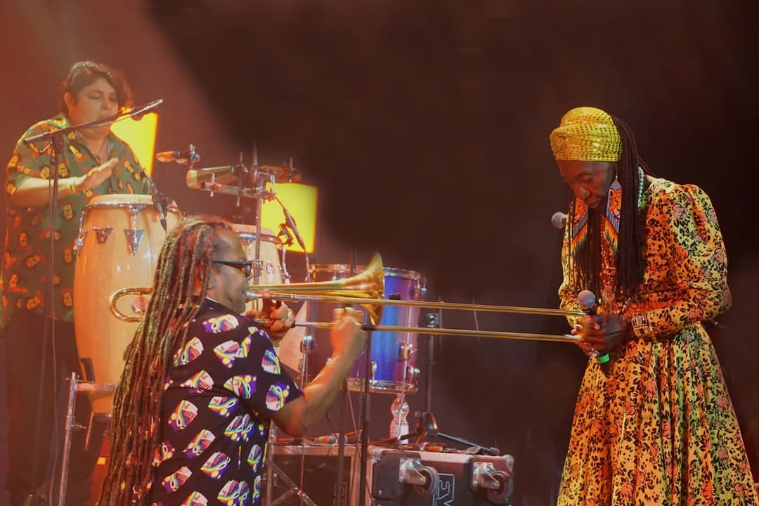 Lia de Itamaracá, de turbante e vestido estampado, canta no palco ao lado de músicos com tambor e trombone
