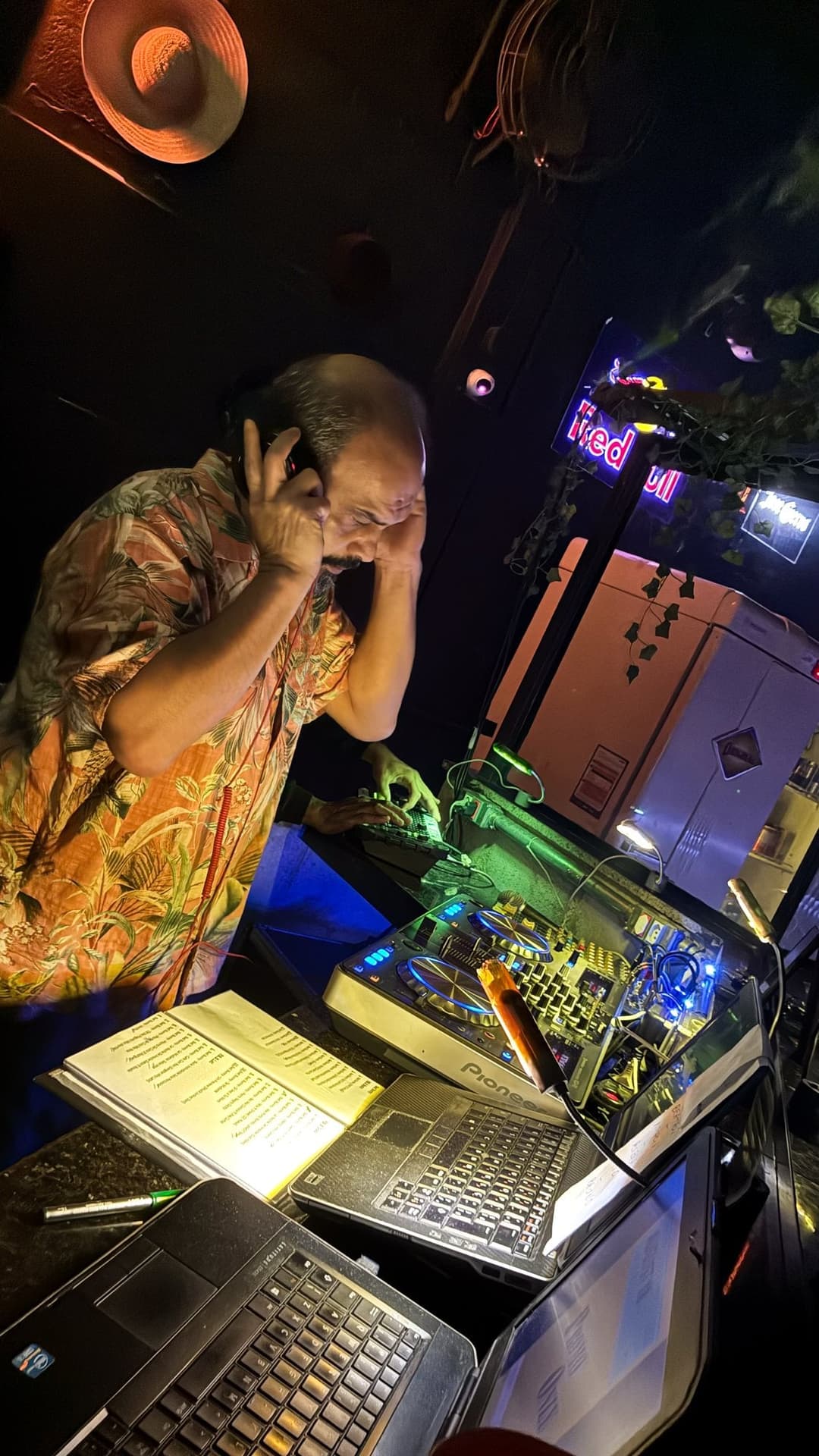 DJ de camisa estampada na cabine, com as mãos nos fones, diante da controladora e de notebooks