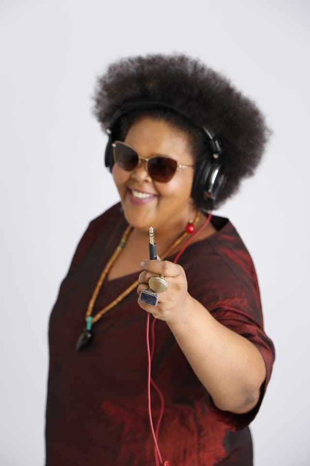 DJ Black Josie sorri de fones de ouvido e óculos escuros, segurando um cabo de áudio em direção à câmera