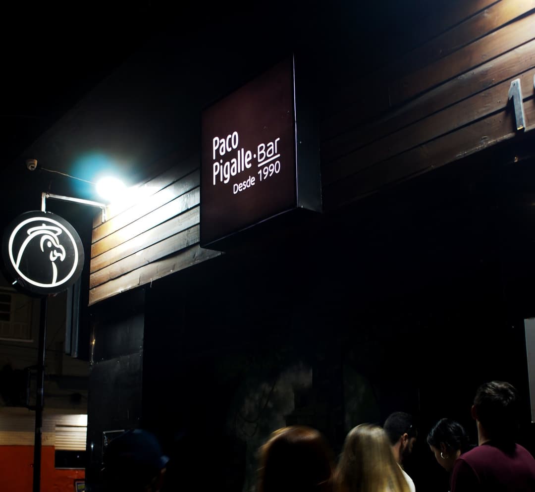 Fachada do Bar Paco Pigalle à noite, com a placa "Paco Pigalle Bar, desde 1990" iluminada e pessoas na entrada