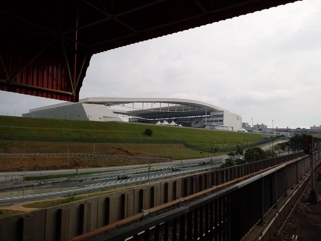 Estádio de fachada branca visto de uma passarela elevada, com gramado e trilhos em primeiro plano